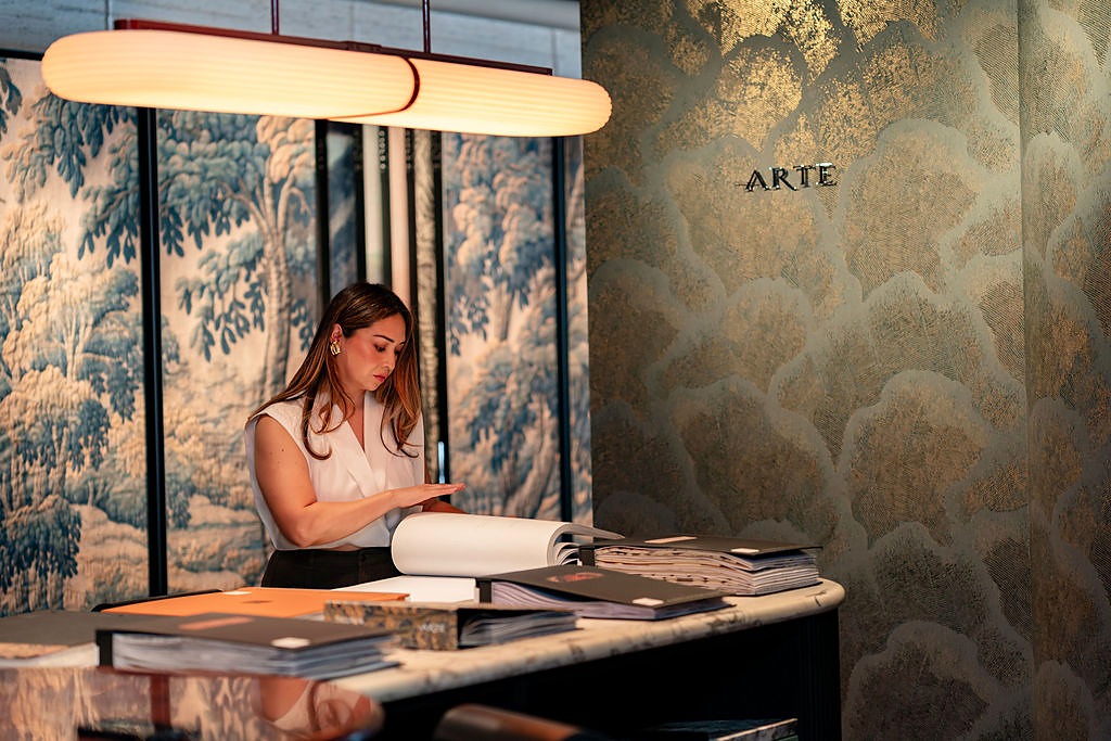 Gisele Cano of The Style Maison paging through wallcovering books in front of an Arte wallcovering in a design showroom